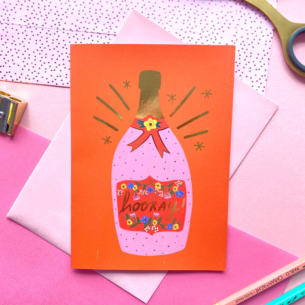 An orange greeting card featuring a stylized pink champagne bottle with the word 'hookey' in script, styled in a flat lay with pink paper, scissors, and colored pencils. Hooray congratulations greeting card