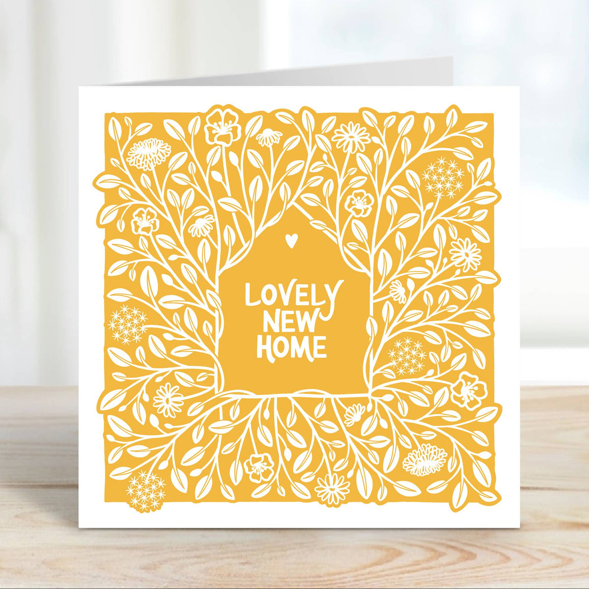A square greeting card with a white border and a yellow floral tree design, standing on a light wooden surface. The center of the card reads 'LOVELY NEW HOME' in white text.