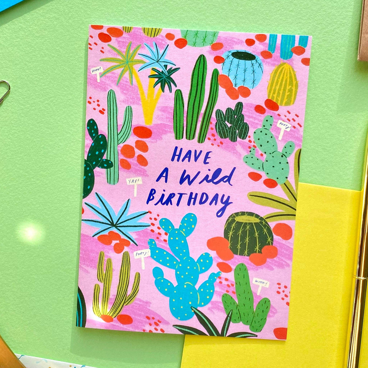 A colorful birthday card with a pink background and a cactus pattern, reading 'HAVE A WILD BIRTHDAY' in blue text. The card is styled in a flat-lay arrangement on a light green surface with gold scissors, a yellow paper sheet, a gold pen, and other stationery props.