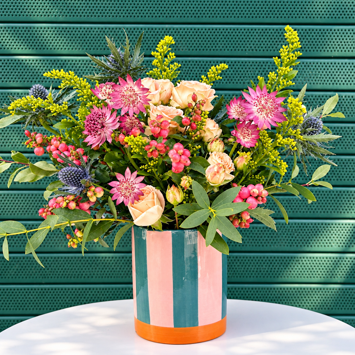 A vibrant floral arrangement in a striped ceramic pot, set on a white table against a green slatted wall.