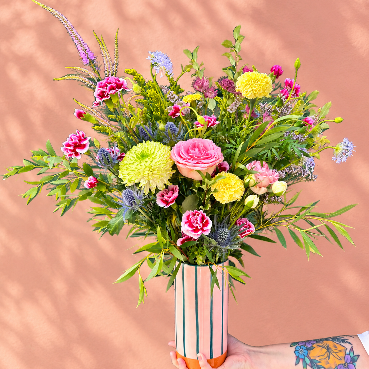 A vibrant, multicolored wildflower bouquet displayed in a colourful striped vase that is pink blue and orange held by a hand with a floral tattoo, set against a warm terracotta background with soft leaf shadows.