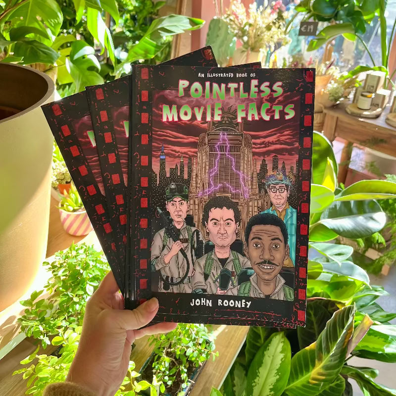 Book titled 'Pointless Movie Facts' by John Rooney held in front of green plants. Funny illustrated comic perfect gift for movie film fans