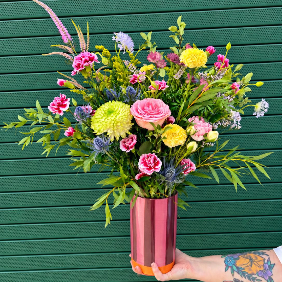 A vibrant floral arrangement in a ceramic pink and red stripe cylindrical vase held by a tattooed arm against a dark green slatted background.