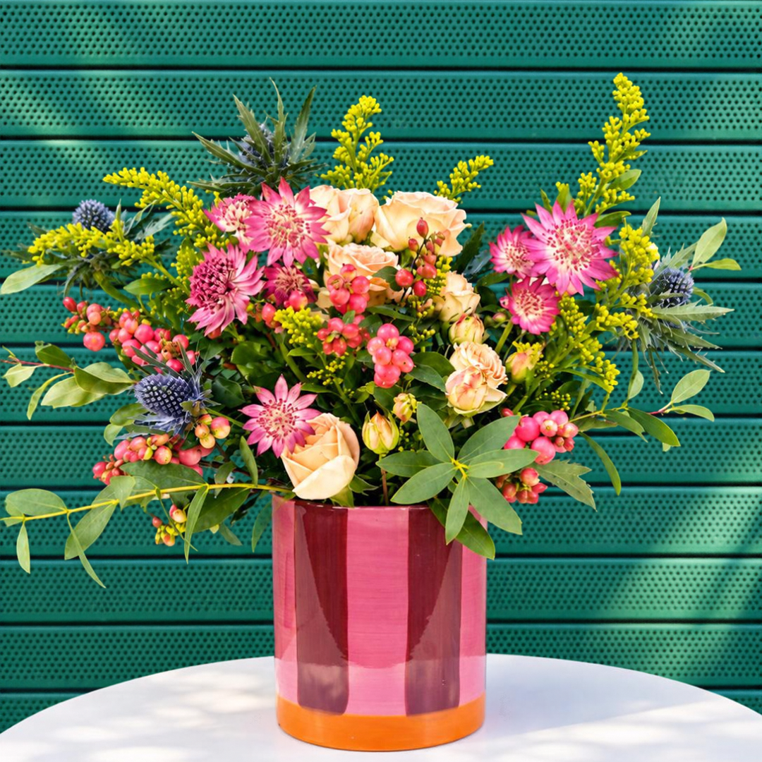 A vibrant floral arrangement in a striped pink and orange cylindrical vase, set on a white table against a green perforated metal wall.